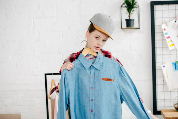 Teenager kid in cap holding hanger with blue shirt at home - Foto, imagen