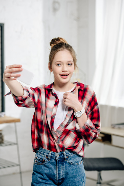 Vidám tinédzser gyerek gazdaság okostelefon és figyelembe selfie hüvelykujjával fel otthon - Fotó, kép