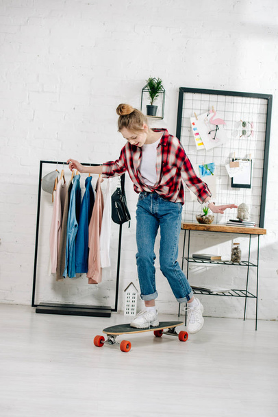 Full length view of teenager in jeans and checkered shirt standing on longboard in bedroom - Photo, Image