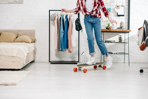 Cropped view of teenager in jeans and checkered shirt standing on longboard in bedroom - Photo, Image