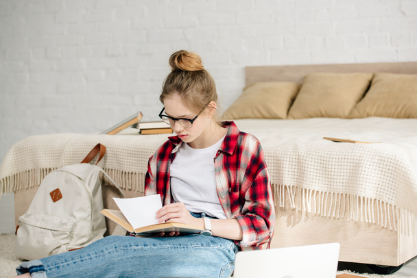Concentrated teenager in glasses and checkered shirt reading book near bed - 写真・画像