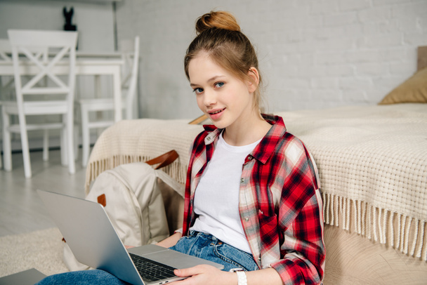 Teenage kid in checkered shirt using laptop while sitting on carpet - Zdjęcie, obraz