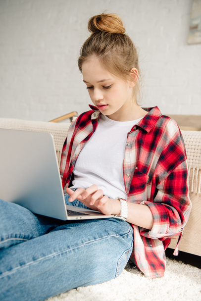 Teenage kid in checkered shirt using laptop while sitting on carpet - Foto, imagen