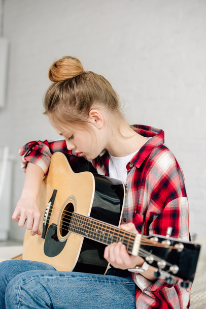 Concentrated teenager in checkered shirt playing acoustic guitar at home - Photo, Image