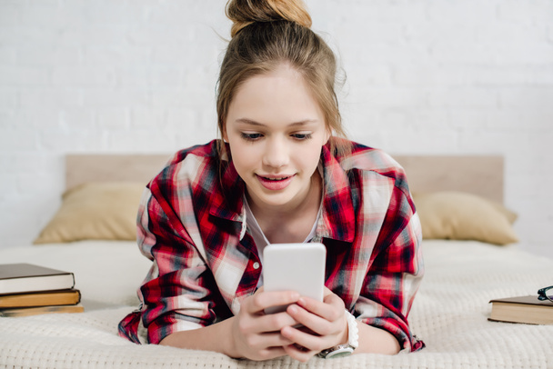 Adolescente sonriente con camisa a cuadros acostada en la cama y usando un teléfono inteligente
 - Foto, imagen