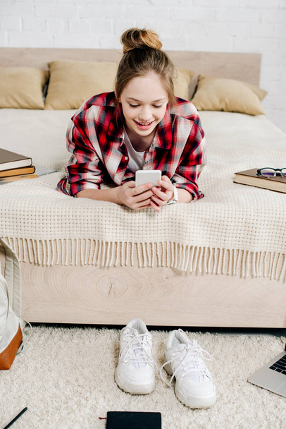 Smiling teenager in checkered shirt lying on bed and using smartphone - Valokuva, kuva