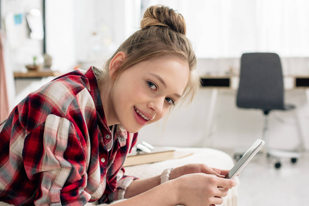 Adolescente sonriente con camisa a cuadros acostada en la cama y usando un teléfono inteligente
 - Foto, imagen