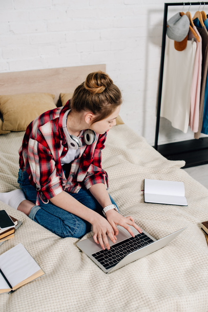 Teenager se sluchátky při psaní na klávesnici laptopu při sezení na posteli - Fotografie, Obrázek