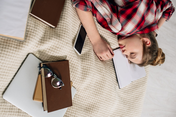 Teenager in checkered shirt sleeping on bed with books and smartphone - Photo, Image