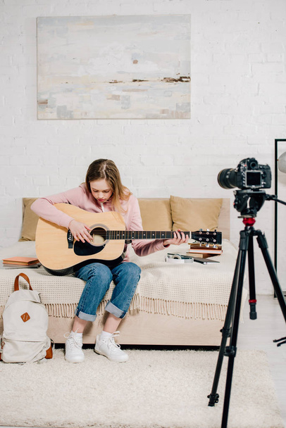 Εφηβικό blogger κάθεται στο κρεβάτι και παίζοντας ακουστική κιθάρα μπροστά από την κάμερα - Φωτογραφία, εικόνα