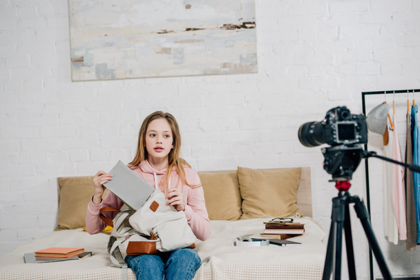 Teenager kid sitting on bed with books and backpack in front of video camera - Photo, Image