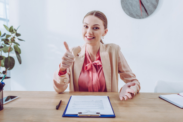 cheerful recruiter showing thumb up while sitting near clipboard and looking at camera - Photo, Image