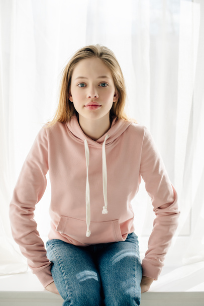 Frontansicht eines Teenagers im rosafarbenen Kapuzenpulli, der auf der Fensterbank sitzt und in die Kamera blickt - Foto, Bild