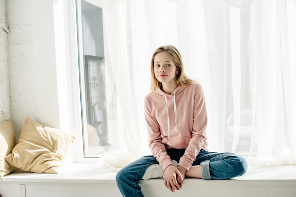 Pensive teenage kid in jeans sitting on window sill and looking away - Photo, Image