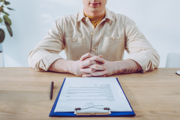 cropped view of man sitting with clenched hands near clipboard and pen  - Fotografie, Obrázek