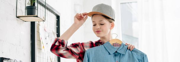 Panoramic shot of teenager in cap holding hanger with blue shirt - Fotografie, Obrázek