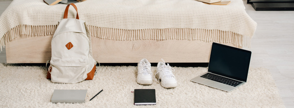 Tiro panorâmico de tênis branco, laptop com tela em branco e mochila no tapete
 - Foto, Imagem