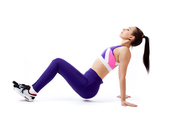 Молодая женщина тренер в спортивном фиолетовый короткий топ и тренажерный зал делает и растягивает ноги, делая упражнения на белом изолированном фоне в студии
 - Фото, изображение