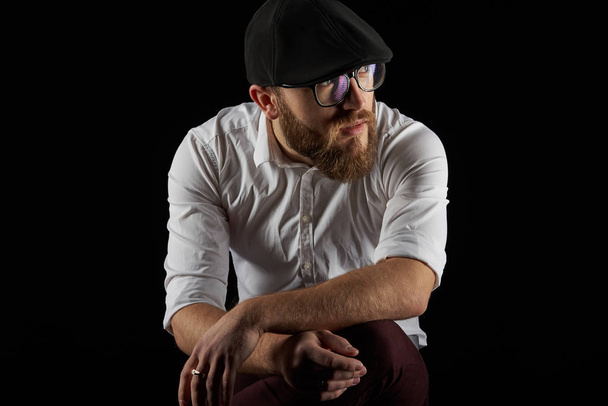 jovem muito barbudo homem em óculos com uma barba e um bigode em um boné preto e uma camisa branca sentada em sua cadeira encostada ao joelho, um fundo escuro estúdio
 - Foto, Imagem