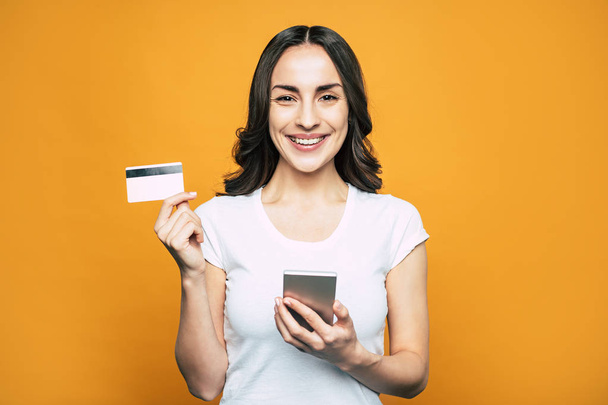 Check card. Bodacious girl with hazel eyes, slightly curled hair and such a brilliant smile in front of flamboyant orange background is holding her phone and a bank card. - Foto, afbeelding
