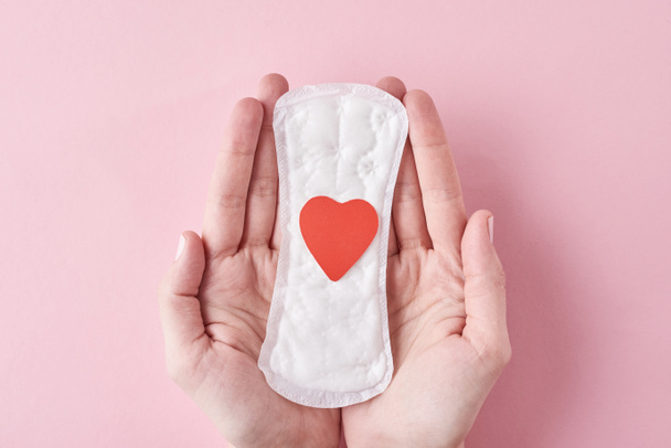 Woman hands hold sanitary pad with red heart on a pink background - Foto, afbeelding