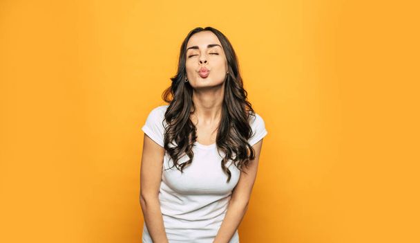 Waving a kiss. An air-kiss made by eupeptic girl with long dark curly hair, glowing tanned skin and nice-looking body in front of flame-orange background. - Photo, image
