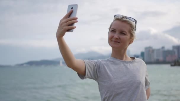 Красива струнка жінка з косичкою блондинка в сірій футболці, що стоїть на мосту і робить селфі на мобільному телефоні над фоном моря і міста. Дівчина торкається екрана
 - Кадри, відео