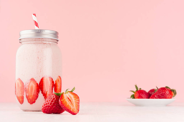 Light dairy strawberry drink in trendy jar with ripe pieces berry on saucer, red striped straw, silver lid on gentle pink background and white wood table. - Foto, afbeelding