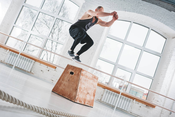 Dynamic shot of fit athlete jumping over cross box in fitness club. Strong man doing jump exercises in gym - 写真・画像