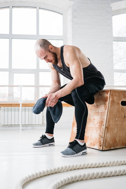 Tired strong man sitting on box at gym. Perspiring fit sportsman resting after hard cross workout in light hall - Foto, Imagen