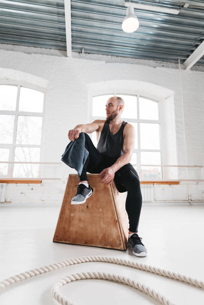 Perspiring male athlete resting after intense cross workout. Tired sportsman relaxing on wooden box in fitness gym - Foto, Imagen