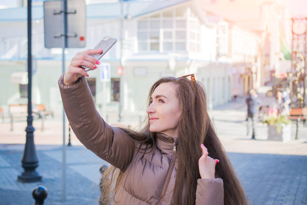 Beautiful young brunette with long hair taking smiling selfi in the european city street - Foto, Imagem