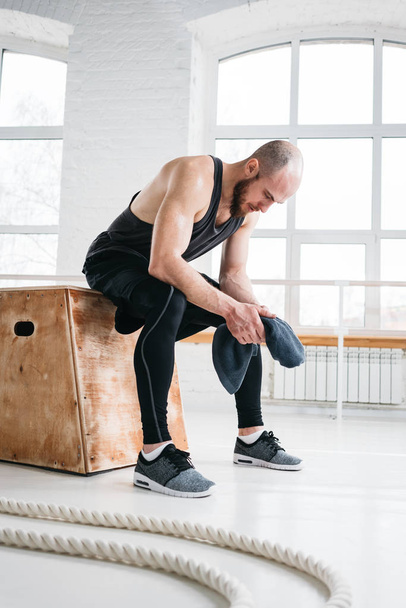 Professional workout athlete resting after cross work session in fitness gym. Male sportsman sitting on wooden box and hold towel in hand. Vertical - Photo, Image