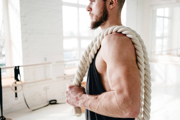 Handsome muscular man holding on shoulder rope workout in light gym. Portrait of fitness man with battle ropes at cross gym - Φωτογραφία, εικόνα