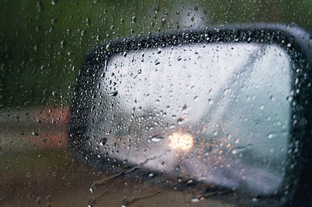 Rear-view mirror with water drops and car with headlights. Low visibility driving. Selective focus, shallow DOF - Фото, изображение