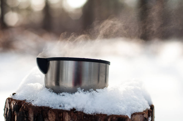 a mug of hot tea stands on a snow-covered stump in the winter, cold, pine forest - Photo, image
