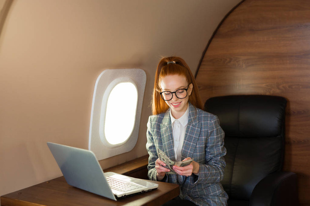 beautiful young girl with red hair sits at a table with a laptop in the cabin of the plane with dollars in their hands - Foto, imagen