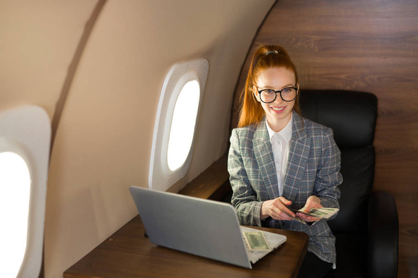 beautiful young girl with red hair sits at a table with a laptop in the cabin of the plane with dollars in their hands - Фото, изображение