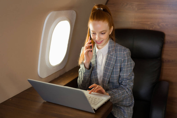 beautiful young girl with red hair sits at a table with a laptop in the cabin of a private jet - Valokuva, kuva