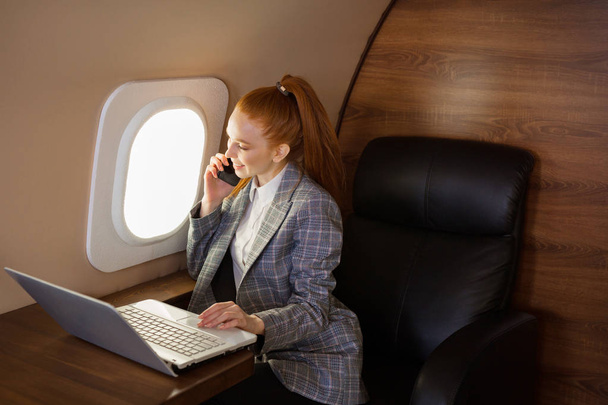 beautiful young girl with red hair sits at a table with a laptop in the cabin of a private jet - Foto, Bild
