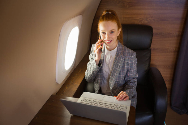 beautiful young girl with red hair sits at a table with a laptop in the cabin of a private jet - Foto, afbeelding