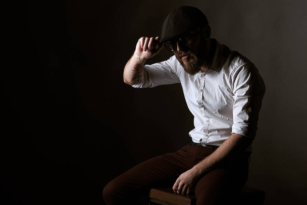 young pretty man in the glasses of fight and mustache in a black cap and white shirt I fix my cap, a dark studio background - Fotografie, Obrázek