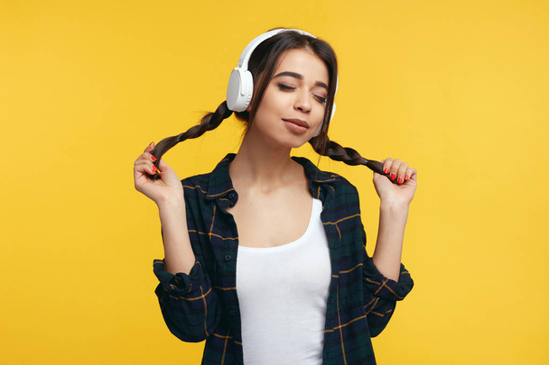 Happy female teenager enjoy sound of music, holds her pony tails, closes eyes from pleasure, listens song and feels glad - Photo, Image