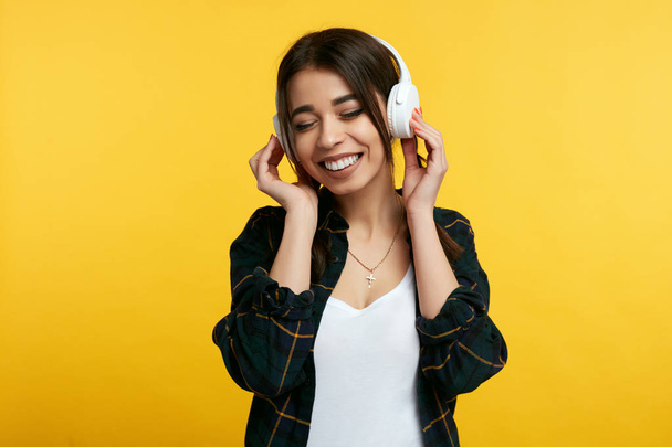 fröhliches junges Mädchen genießt den Klang der Musik, hält die Hände auf Kopfhörer, schließt vor Vergnügen die Augen, isoliert vor gelbem Hintergrund - Foto, Bild