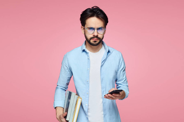 Joven estudiante barbudo confiado sosteniendo libros y teléfono móvil, se ve serio, mientras está de pie aislado sobre el telón de fondo rosa
 - Foto, Imagen