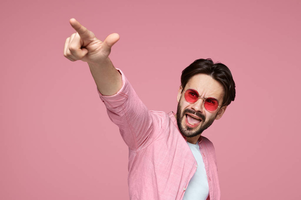 Handsome man in pink shirt pointing finger at camera, screaming and standing over pink background. - Foto, Imagen