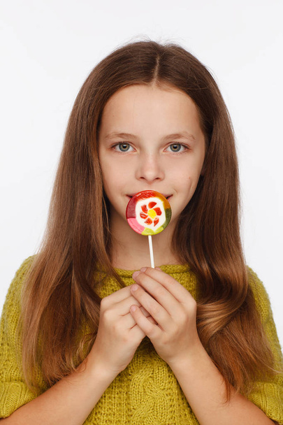 Emotional portrait of an eight-year girl in a yellow-green sweater and with a lollipop in her hands - Фото, изображение