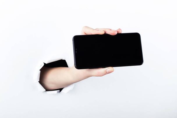 Female hand out of the hole in the paperman, holding a smartphone with a black screen. Isolate on white background. - Foto, immagini