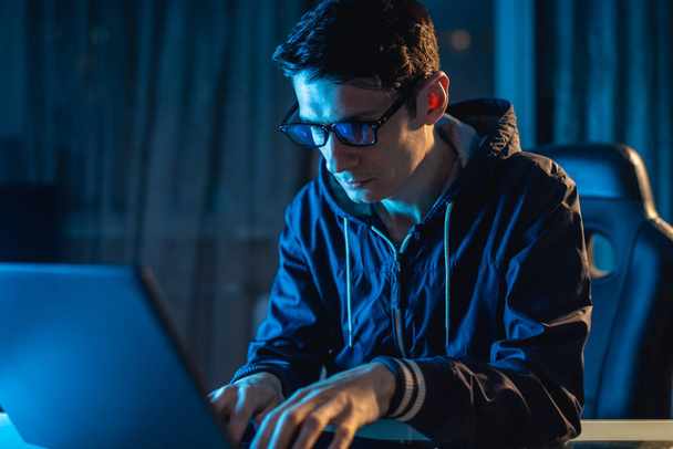 A young man with glasses working on a laptop at night. Designer or system administrator stayed late at work. - Foto, imagen