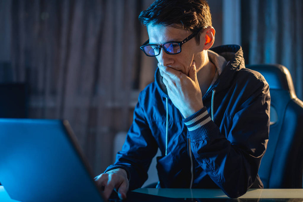 Young man with glasses working on a laptop at night. Freelancer is working late on a project at home. - Photo, Image